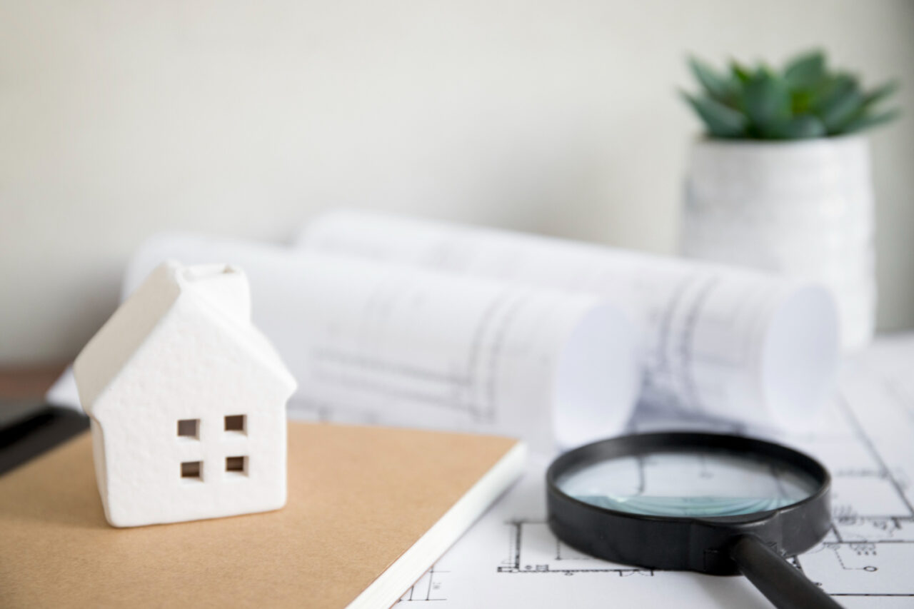 Cahier, loupe et documents posés près d’une maison symbolisant le diagnostic, la préparation et la vérification avant l’achat d’un bien immobilier